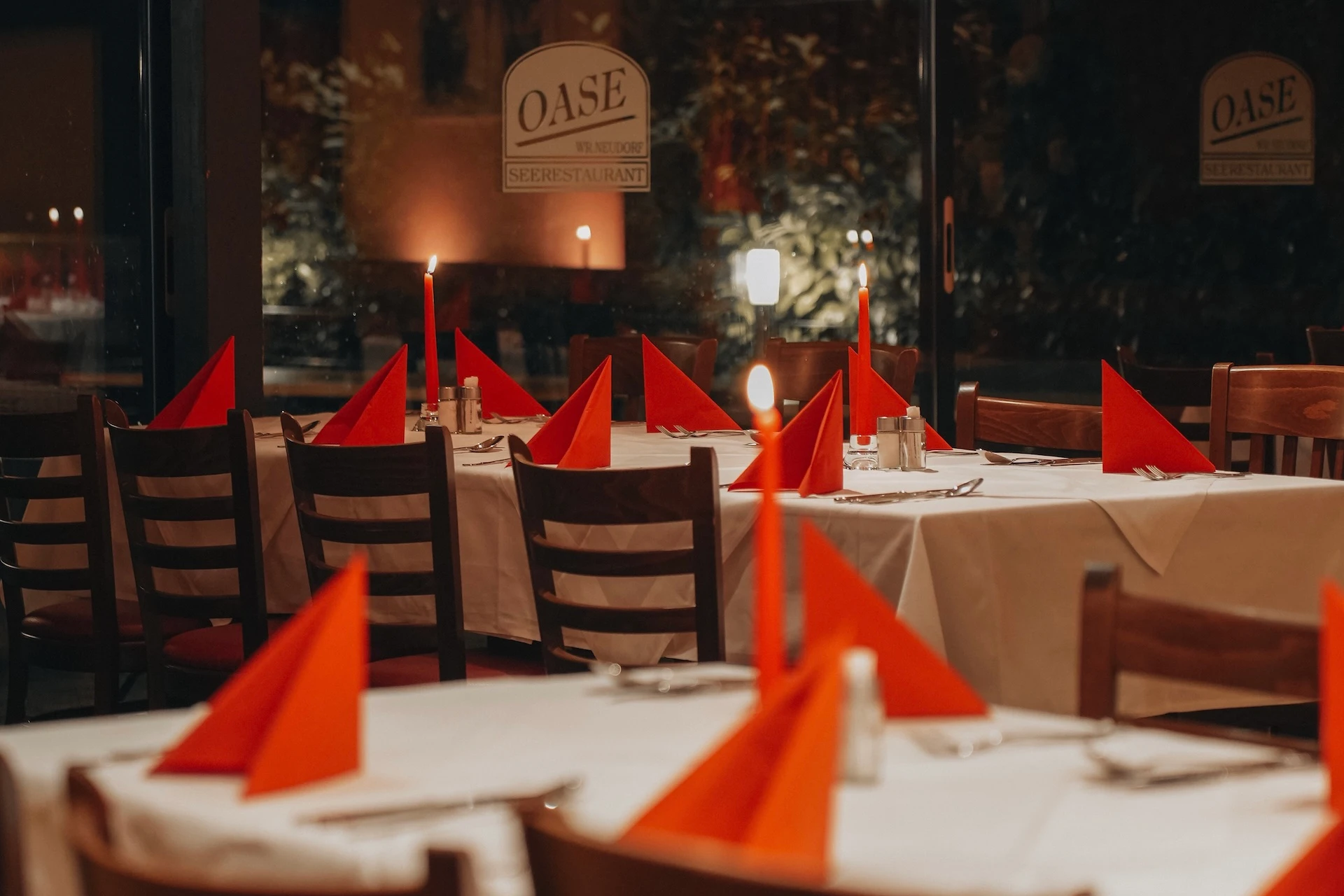Elegante Restauranttische mit weißen Tischdecken, gedeckt mit roten, zu Dreiecken gefalteten Servietten und beleuchtet von roten Kerzen, die eine warme, einladende Atmosphäre schaffen. Im Hintergrund ist ein Fenster mit der Aufschrift „OASE Restaurant“ zu sehen.