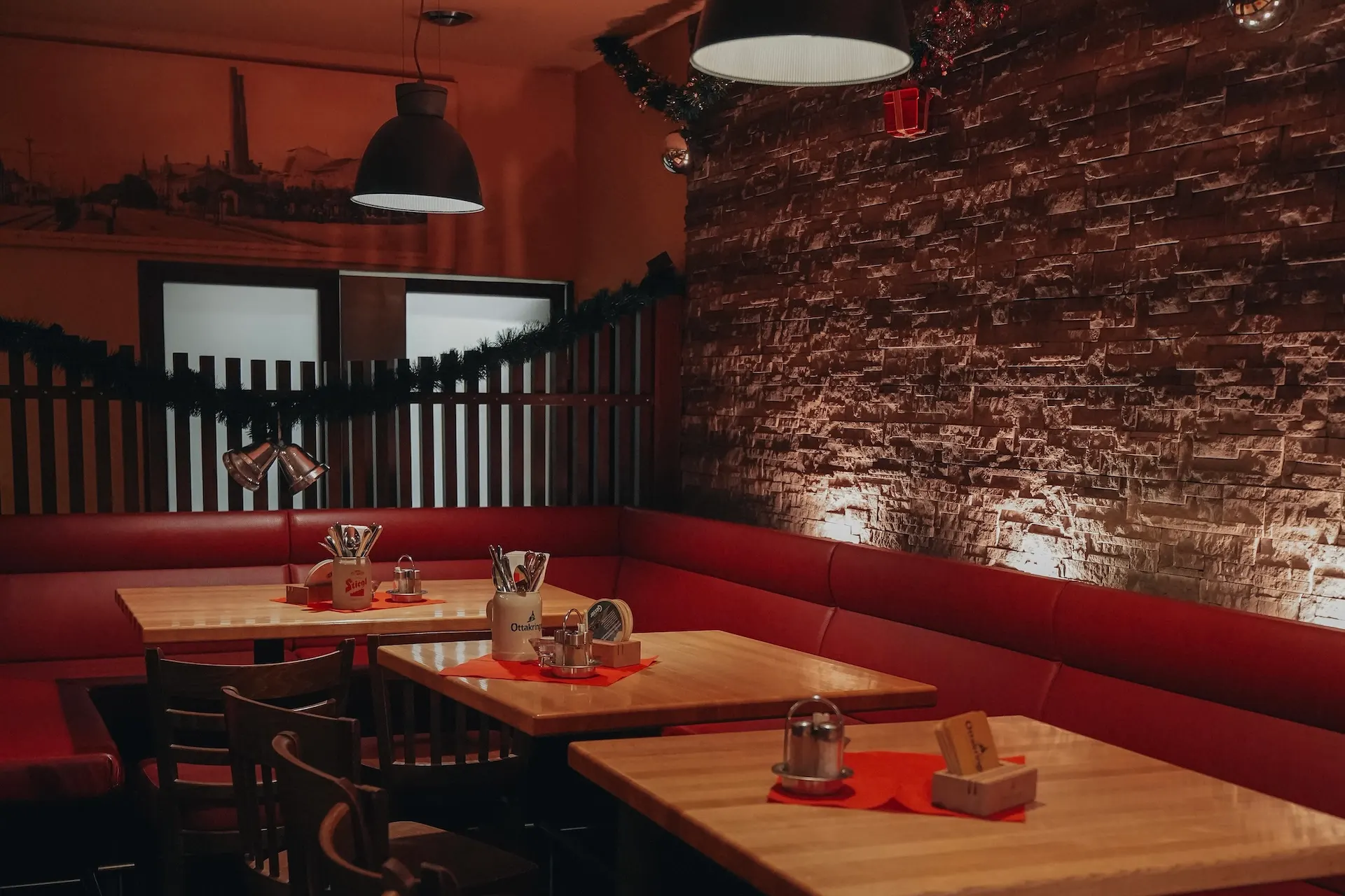 Oase am Teich Gemütliches Restaurantinterieur mit Holztischen, roten gepolsterten Sitzgelegenheiten, einer unverputzten Backsteinwand und warmer Beleuchtung. Auf den Tischen stehen Gewürze und Servietten bereit; eine festliche Girlande schmückt die Wand. Es sind keine Personen zu sehen.