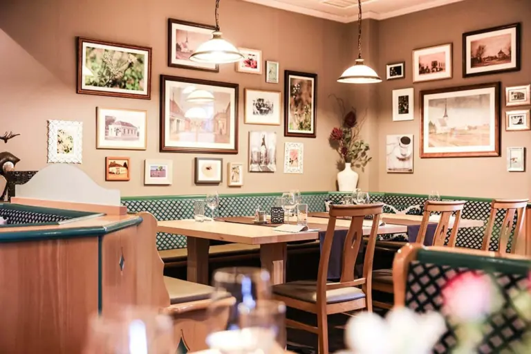 Gemütliches Restaurantinterieur mit Holztischen und -stühlen, Gedecken und einer mit zahlreichen gerahmten Fotos und Kunstwerken dekorierten Wand. Die sanfte Beleuchtung schafft eine warme, einladende Atmosphäre.