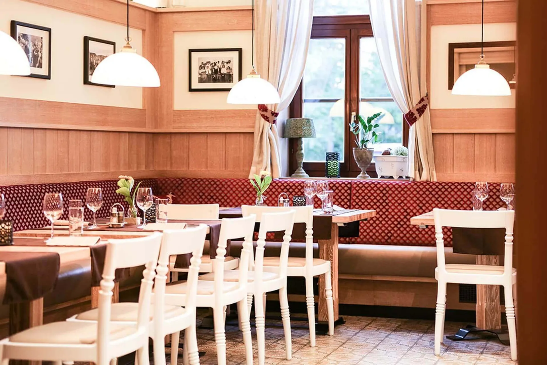 Ein gemütliches Restaurant mit Holzwänden, rot gemusterten, gepolsterten Bänken, weißen Stühlen, gedeckten Tischen mit Gläsern und Servietten, gerahmten Fotos an den Wänden und Sonnenlicht, das durch die vorgehängten Fenster fällt.