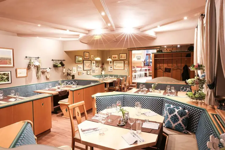 Ein gemütliches Restaurant mit Holztischen und blau gepolsterten Sitzgelegenheiten, die mit Gläsern und Besteck ausgestattet sind. Die Wände sind mit gerahmten Bildern und Pflanzen dekoriert, und die warme Beleuchtung schafft eine einladende Atmosphäre.
