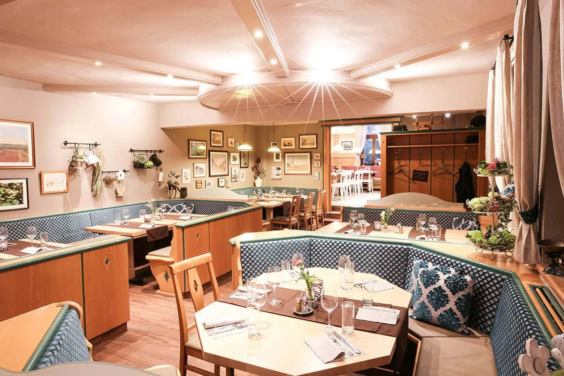 Ein gemütliches Restaurant mit Holztischen und blau gepolsterten Sitzgelegenheiten, die mit Gläsern und Besteck ausgestattet sind. Die Wände sind mit gerahmten Bildern und Pflanzen dekoriert, und die warme Beleuchtung schafft eine einladende Atmosphäre.