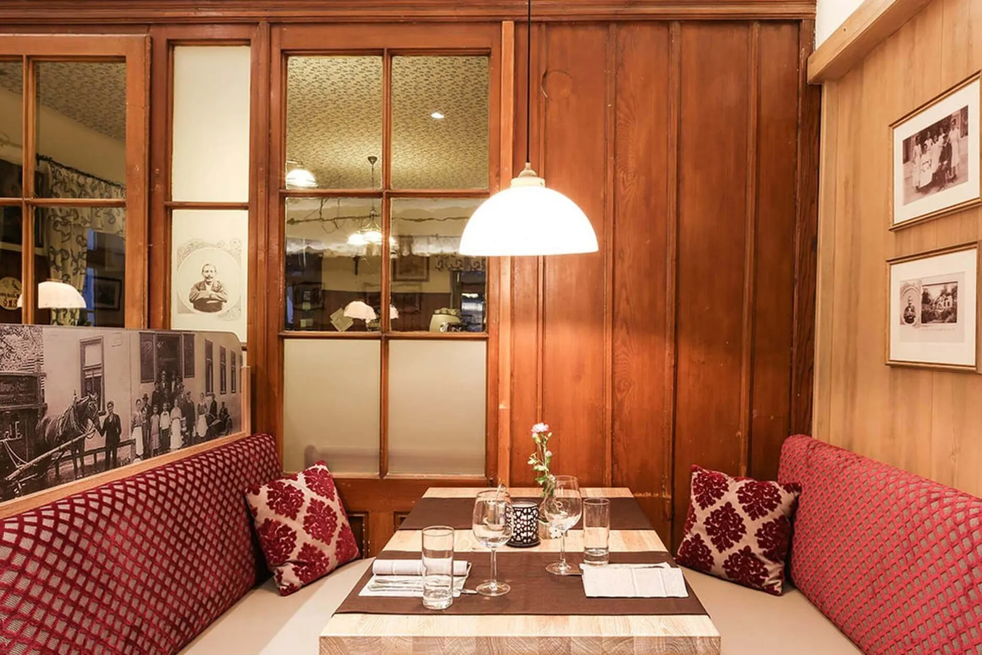 Eine gemütliche Restaurantkabine mit rot gemusterten Kissen, ein Holztisch für vier Personen, darüber eine Hängelampe, holzgetäfelte Wände, gerahmte Bilder und eine kleine Vase mit einer Rose auf dem Tisch.