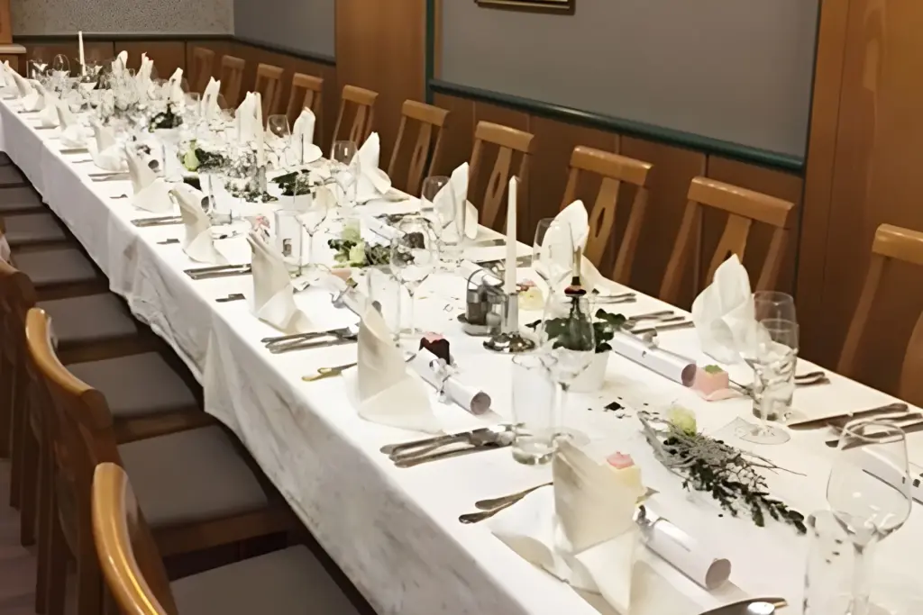 Ein langer Esstisch, gedeckt für eine formelle Veranstaltung mit ordentlich gefalteten weißen Servietten, Besteck, Gläsern, Kerzen und kleinen Blumendekorationen. An beiden Seiten des Tisches sind Holzstühle aufgestellt.