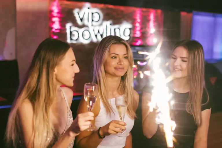 Drei junge Frauen feiern mit Sektgläsern auf einer VIP-Bowlingbahn, eine von ihnen hält eine Wunderkerze in der Hand. Das neonfarbene VIP-Bowling-Schild leuchtet im Hintergrund und schafft eine festliche Atmosphäre.