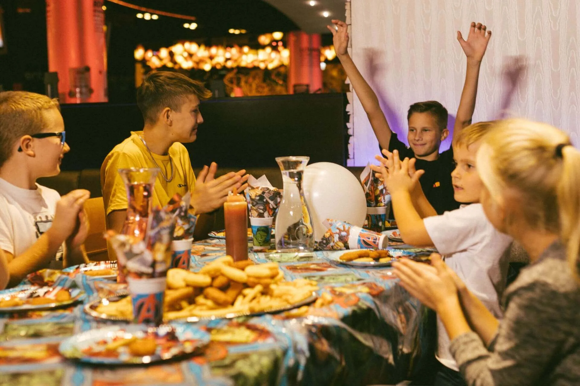 Eine Gruppe von Kindern sitzt um einen mit Snacks und Partydekoration gedeckten Tisch, klatscht und lächelt, während ein Junge aufgeregt die Arme hebt. Die Umgebung wirkt festlich und lebendig, möglicherweise eine Geburtstagsparty.