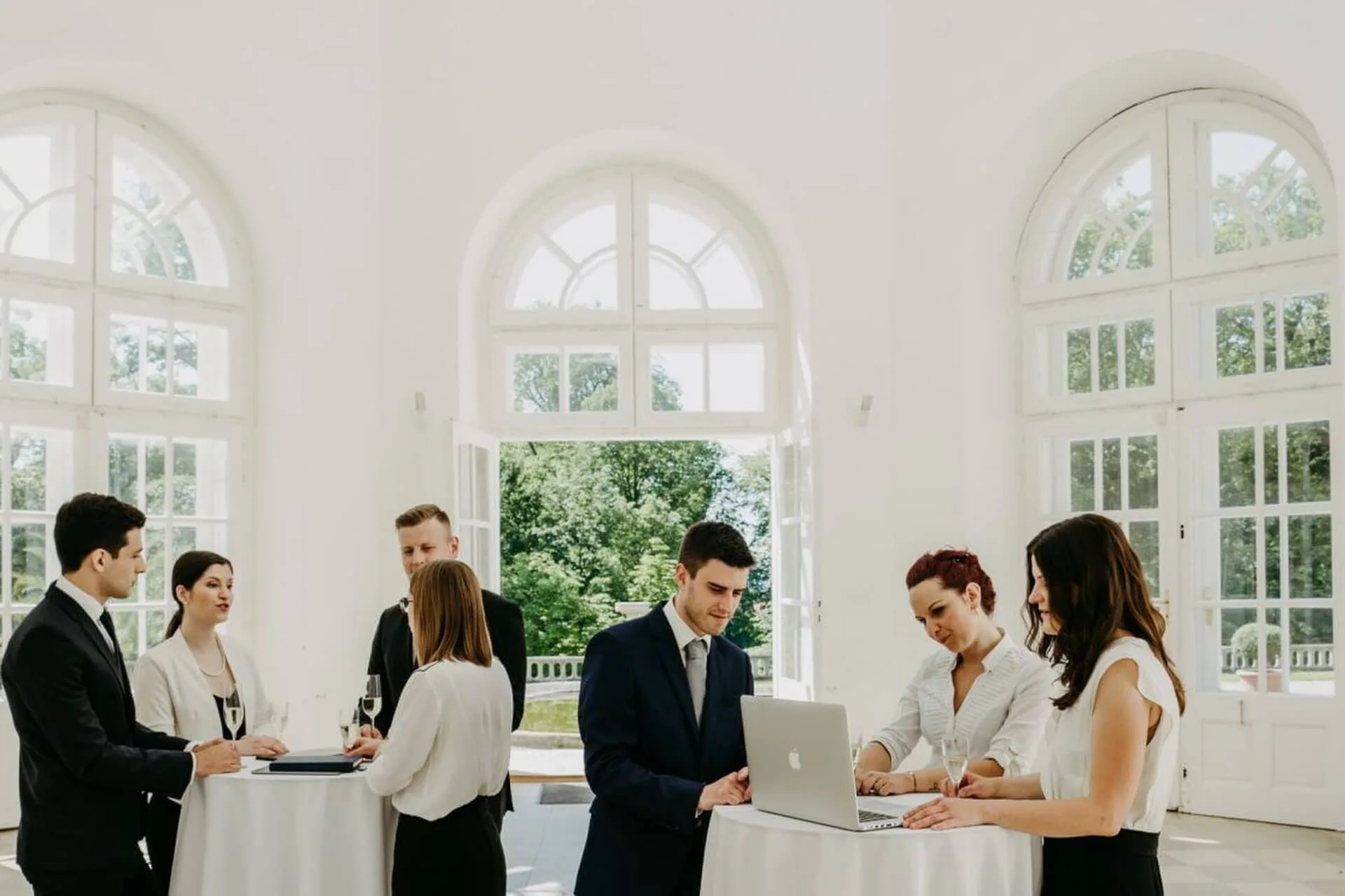 Eine Gruppe professionell gekleideter Menschen steht in einem hellen, eleganten Raum mit hohen Bogenfenstern und unterhält sich. Einige arbeiten an einem runden Tisch an einem Laptop, während andere sich in der Nähe paarweise unterhalten.