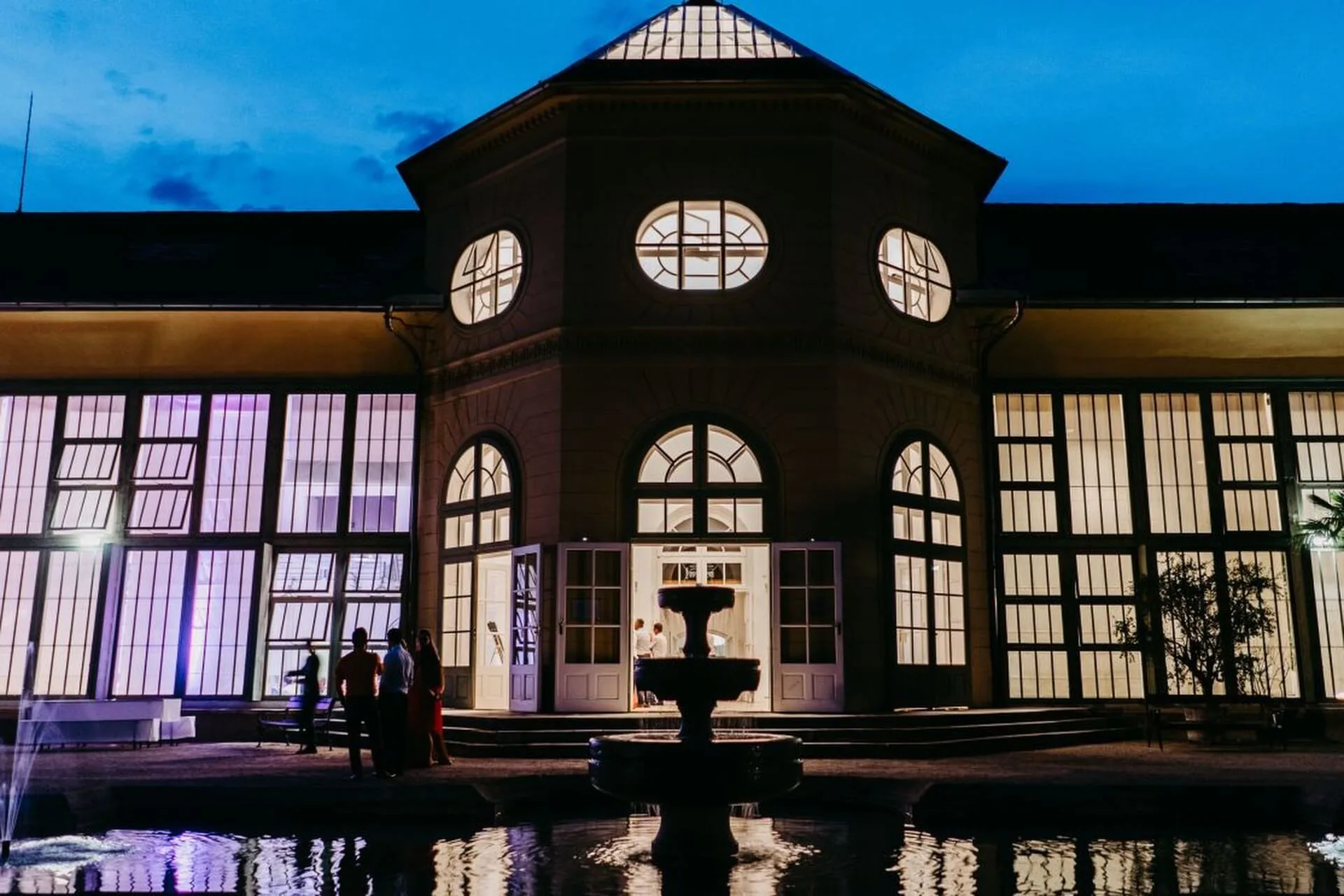 Ein großes, elegantes Gebäude mit hohen, beleuchteten Fenstern in der Abenddämmerung. Menschen stehen in der Nähe des Eingangs und eines zentralen Springbrunnens, der sich in einem Becken im Vordergrund spiegelt.