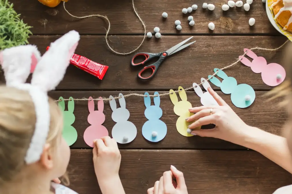 Ein Kind und ein Erwachsener schmücken einen Holztisch mit einer Girlande aus bunten Papierhasen - eine ideale Osterdeko-Bastelaktion. Schere, Schnur, Süßigkeiten und eine rote Klebetube liegen ebenfalls auf dem Tisch.