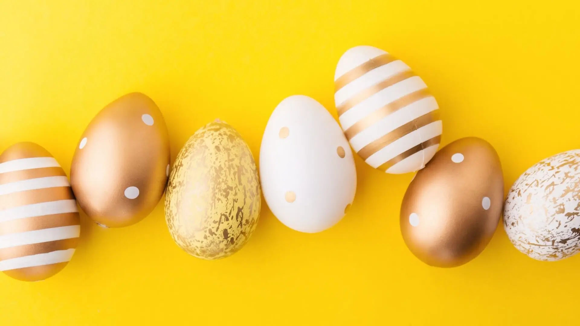 Eine Reihe dekorativer Ostereier mit goldenen, weißen und gold-weißen Mustern, einschließlich Tupfen und Streifen, angeordnet auf einem leuchtend gelben Hintergrund.