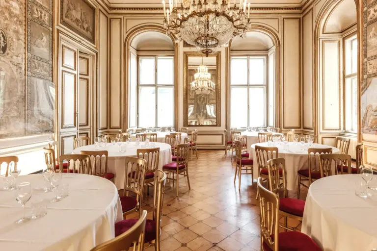 Eleganter Speisesaal mit runden, weiß gedeckten Tischen, goldenen Stühlen mit roten Kissen, großen Fenstern, verzierten Wänden und einem großen Kristalllüster, der sich in einem Wandspiegel spiegelt.