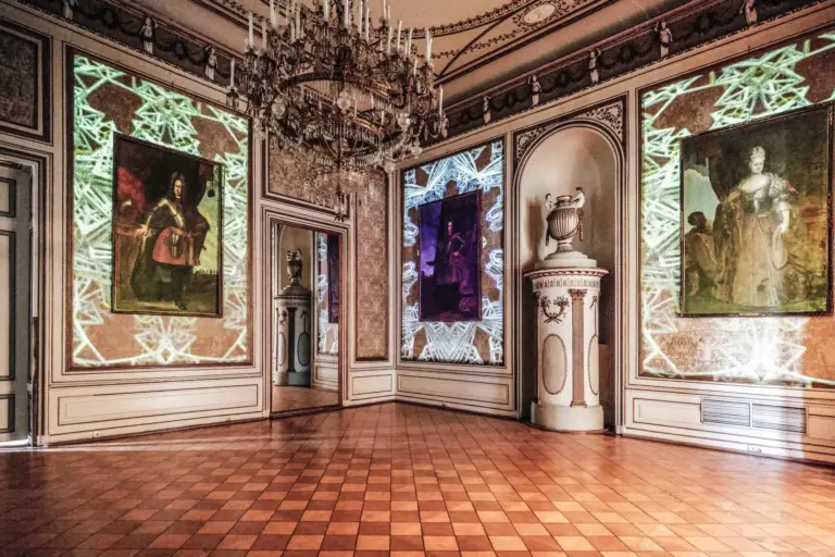 Ein prächtiger, verzierter Raum mit Parkettboden, Kronleuchtern und großen gerahmten Porträts an den Wänden. Dekorative Lichtprojektionen mit geometrischen Mustern erhellen die Gemälde und eleganten architektonischen Details.