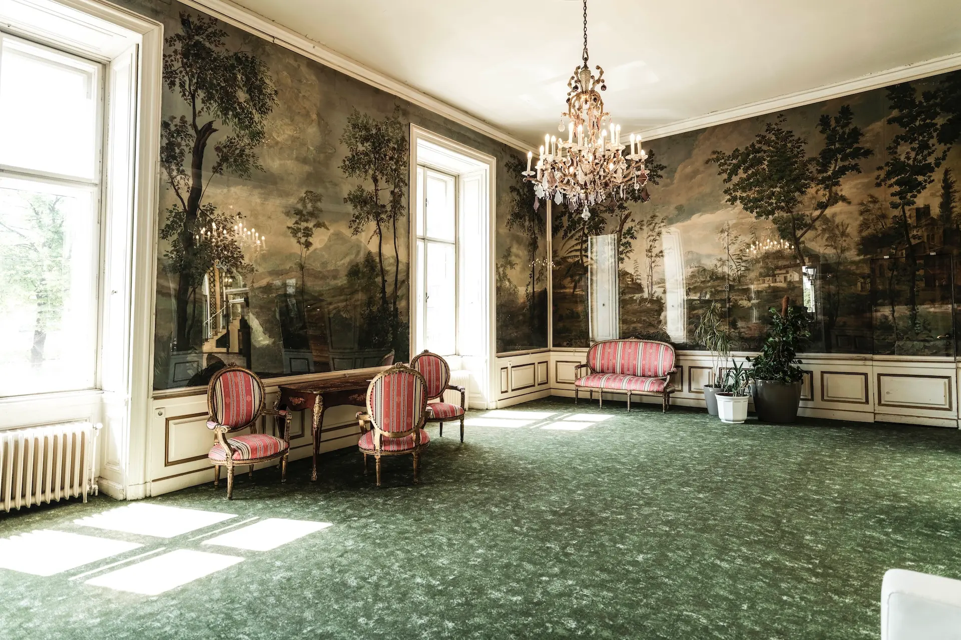 Elegantes Zimmer im Vintage-Stil mit großen Fenstern, grünem Teppich, einem Kronleuchter, einem Holztisch mit verzierten Stühlen, einem rosa gestreiften Sofa, Topfpflanzen und malerischen Wandgemälden, die Bäume und Landschaften darstellen.