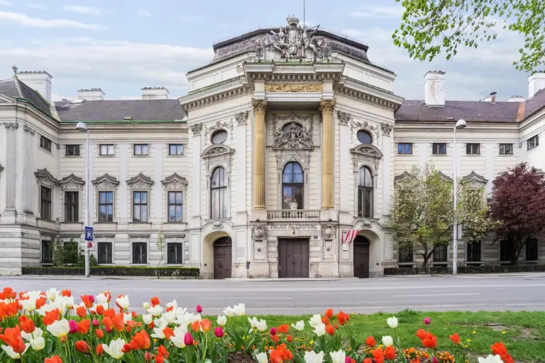 Palais Auersperg Historisches europäisches Gebäude mit verschnörkelten Säulen und Skulpturdetails, umgeben von Bäumen und blühenden roten und weißen Tulpen im Vordergrund, unter einem teilweise bewölkten Himmel.