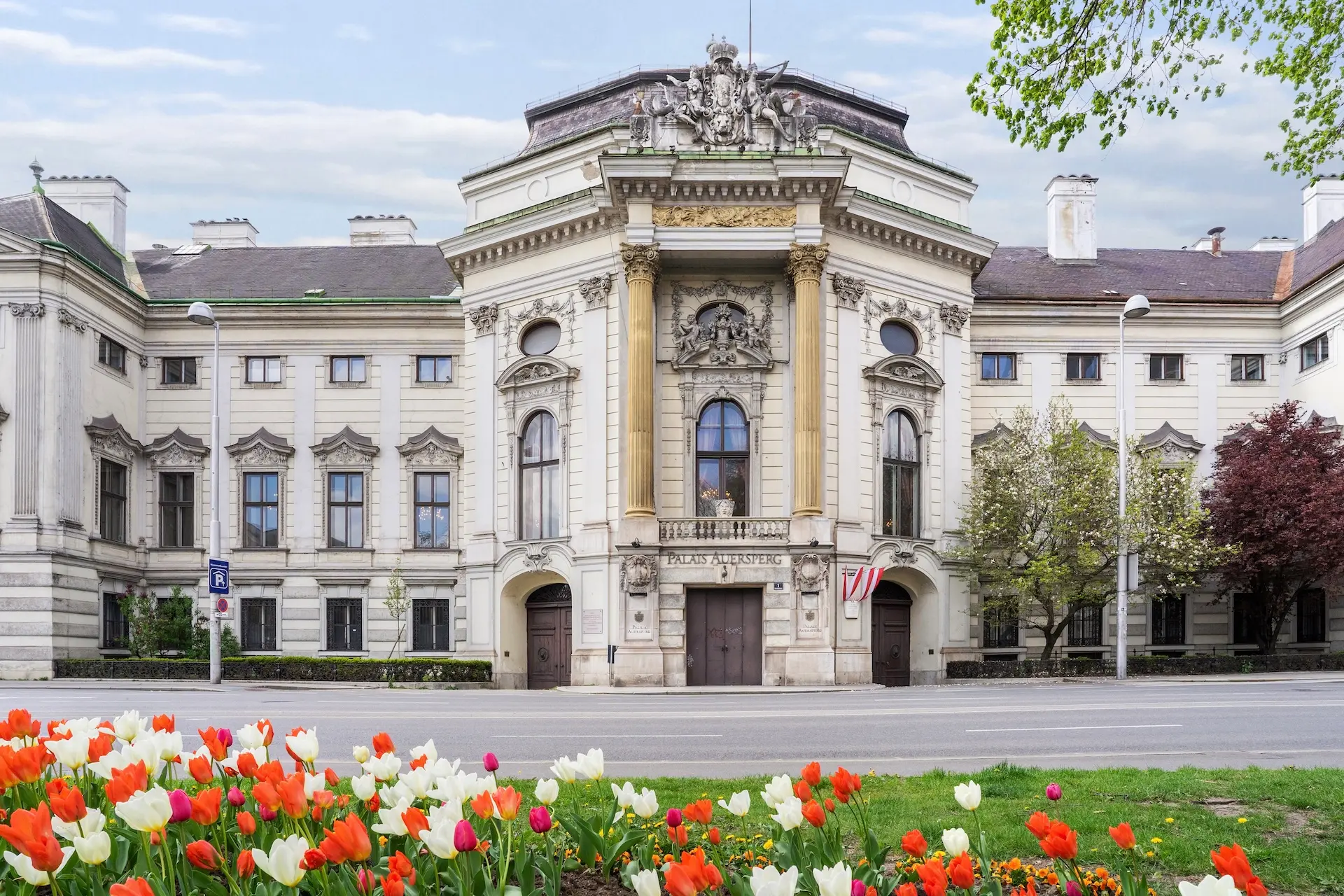 Palais Auersperg Historisches europäisches Gebäude mit verschnörkelten Säulen und Skulpturdetails, umgeben von Bäumen und blühenden roten und weißen Tulpen im Vordergrund, unter einem teilweise bewölkten Himmel.