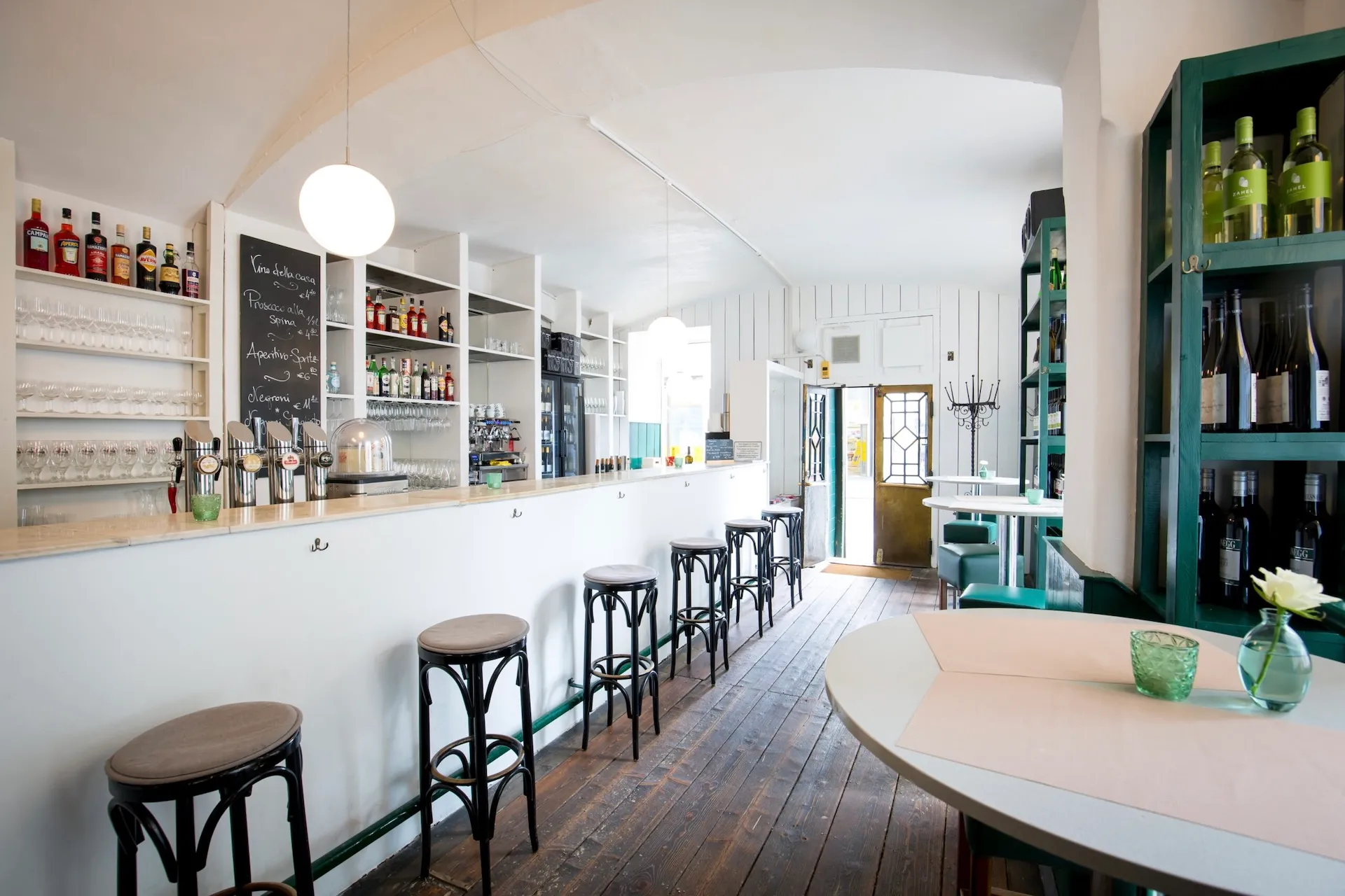 Eine helle, moderne Bar mit weißen Wänden, Holzboden, hohen Barhockern entlang der Theke, Regalen voller Flaschen und einigen runden Tischen mit grünen gepolsterten Stühlen und Blumen in Vasen. Der Raum ist von Tageslicht durchflutet.