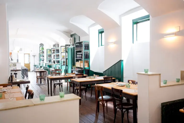 Panigl Trattoria Ein helles, modernes Restaurant mit Holztischen und -stühlen, grünen Zierleisten, Regalen voller Flaschen und Gedecken mit grünen Glasbechern. Durch die großen Fenster strömt Tageslicht herein.