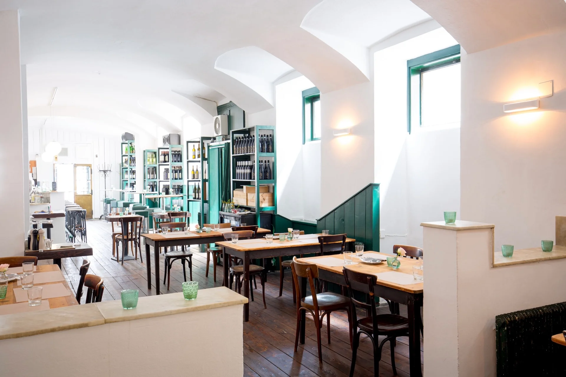 Panigl Trattoria Ein helles, modernes Restaurant mit Holztischen und -stühlen, grünen Zierleisten, Regalen voller Flaschen und Gedecken mit grünen Glasbechern. Durch die großen Fenster strömt Tageslicht herein.