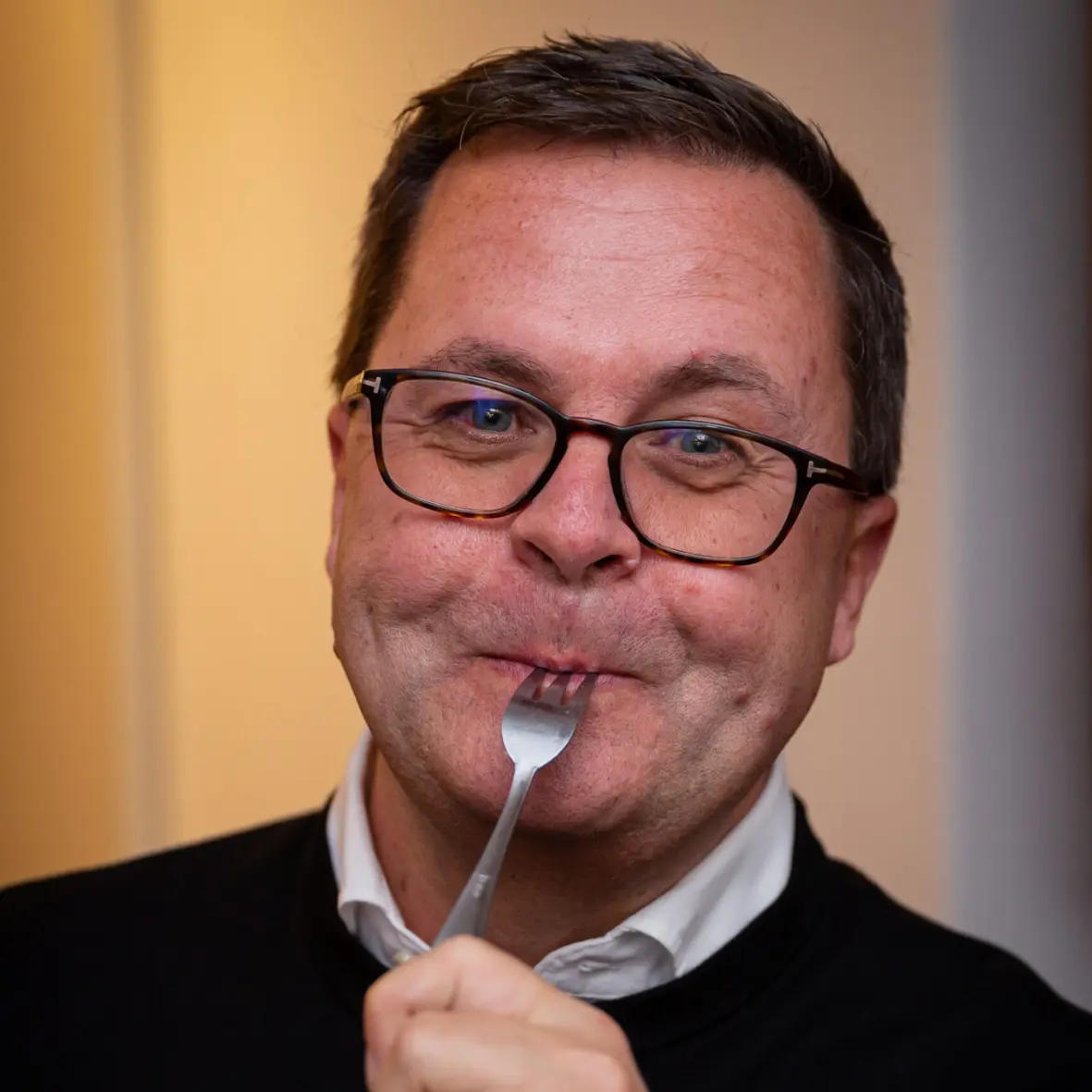 Ein Mann mit Brille und kurzen Haaren lächelt, während er sich eine Gabel an die Lippen führt. Er trägt ein weißes Hemd und einen dunklen Pullover. Der Hintergrund ist sanft beleuchtet und unscharf.