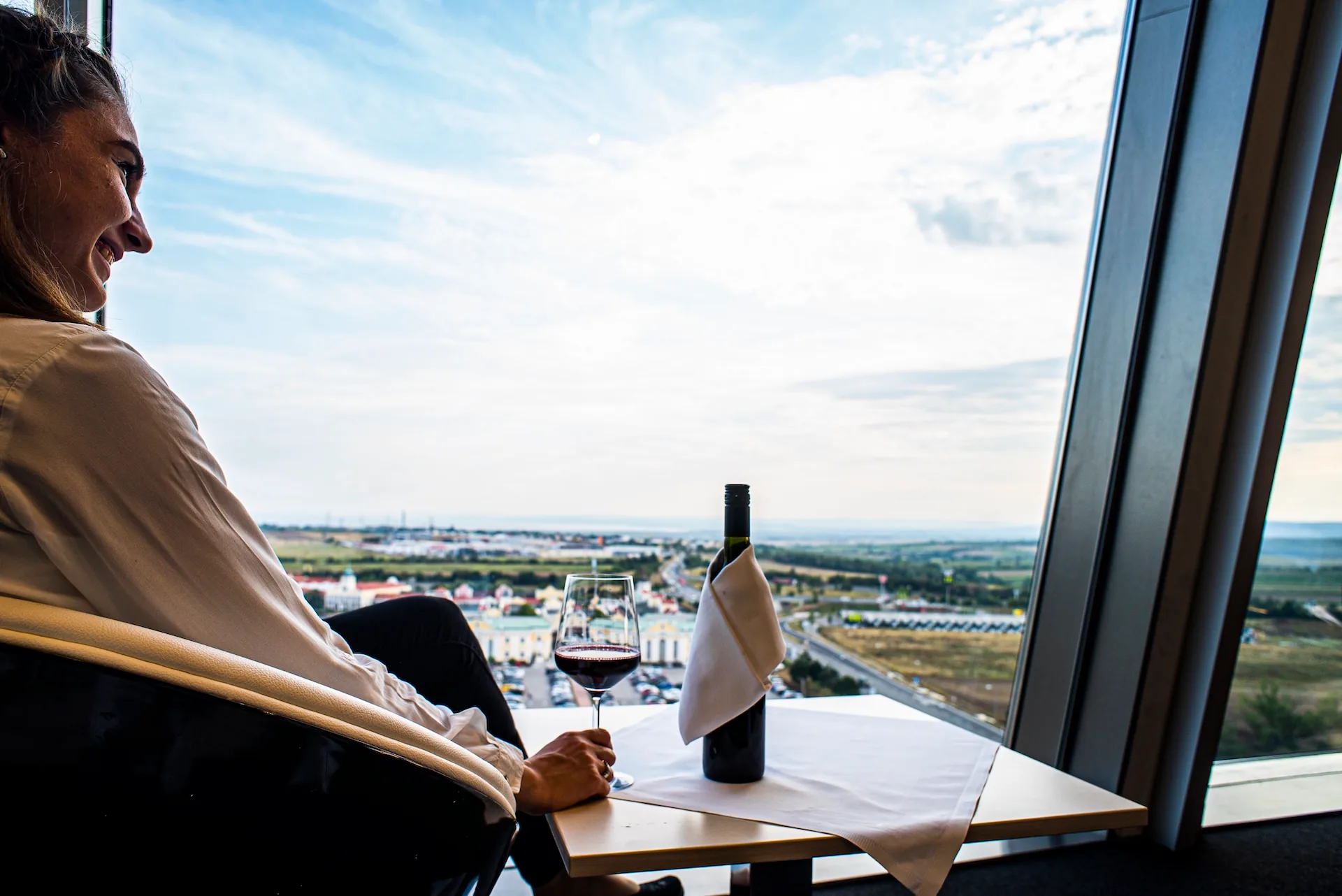 Eine Frau sitzt an einem Fenster mit einem Glas Rotwein und einer Weinflasche auf dem Tisch und blickt auf eine malerische Landschaft mit Feldern und Gebäuden unter einem teilweise bewölkten Himmel.