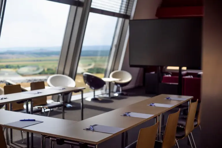 Ein moderner Konferenzraum mit großen schrägen Fenstern, leeren Tischen und Stühlen, die in U-Form angeordnet sind, Klemmbrettern und Stiften auf den Tischen und zwei weißen Stühlen in der Nähe der Fenster mit Blick auf eine malerische Landschaft.