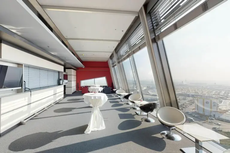Eine moderne Lounge mit raumhohen Fenstern und Blick auf die Stadt, mit weißen Cocktailtischen mit Tüchern und eleganten weißen und schwarzen Stühlen, die entlang des Fensters angeordnet sind.