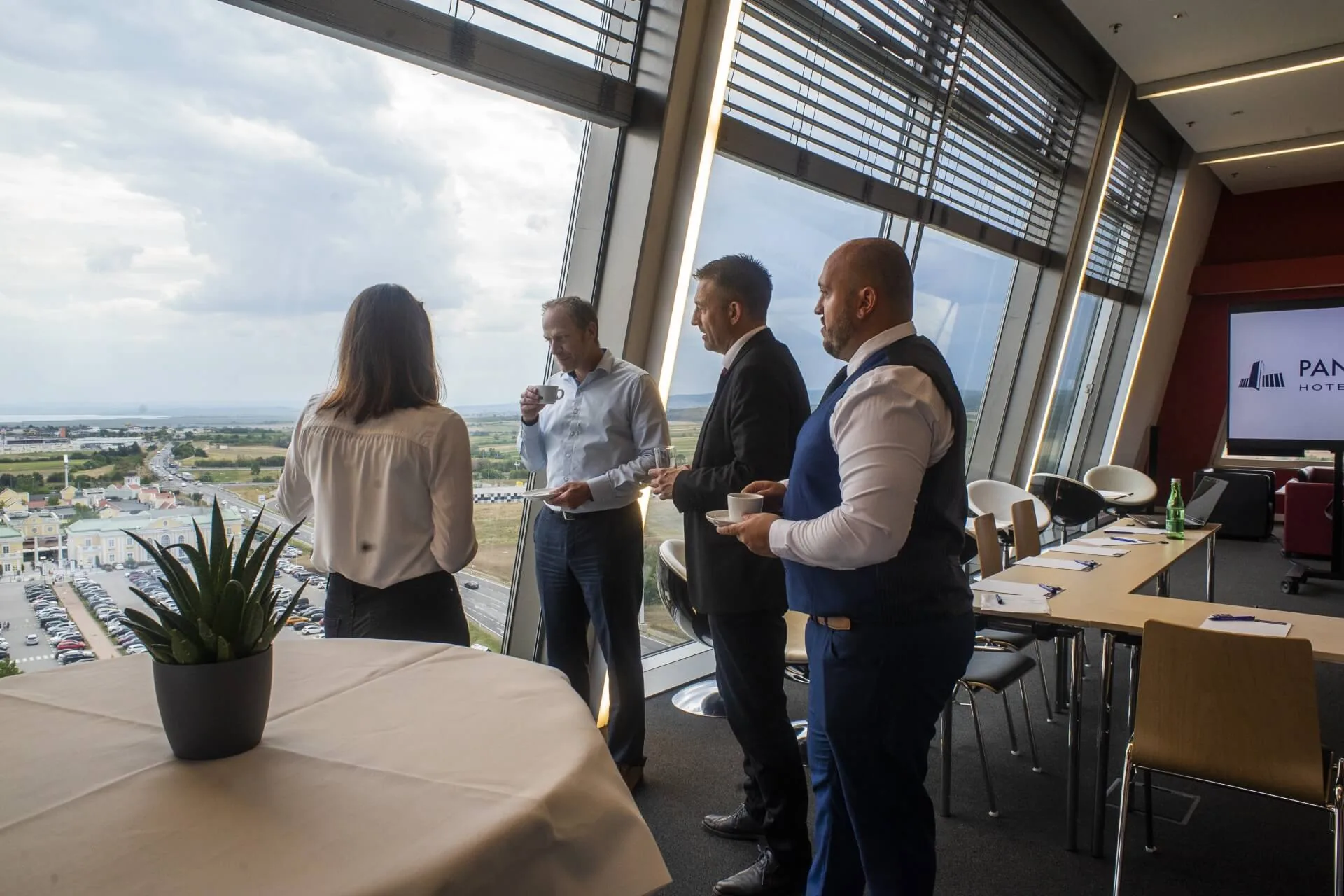 Vier Personen in Geschäftskleidung stehen und unterhalten sich an großen Fenstern in einem modernen Konferenzraum mit Blick auf die Stadt. Ein Tisch mit einer Topfpflanze und eine Reihe von Stühlen sind ebenfalls zu sehen.