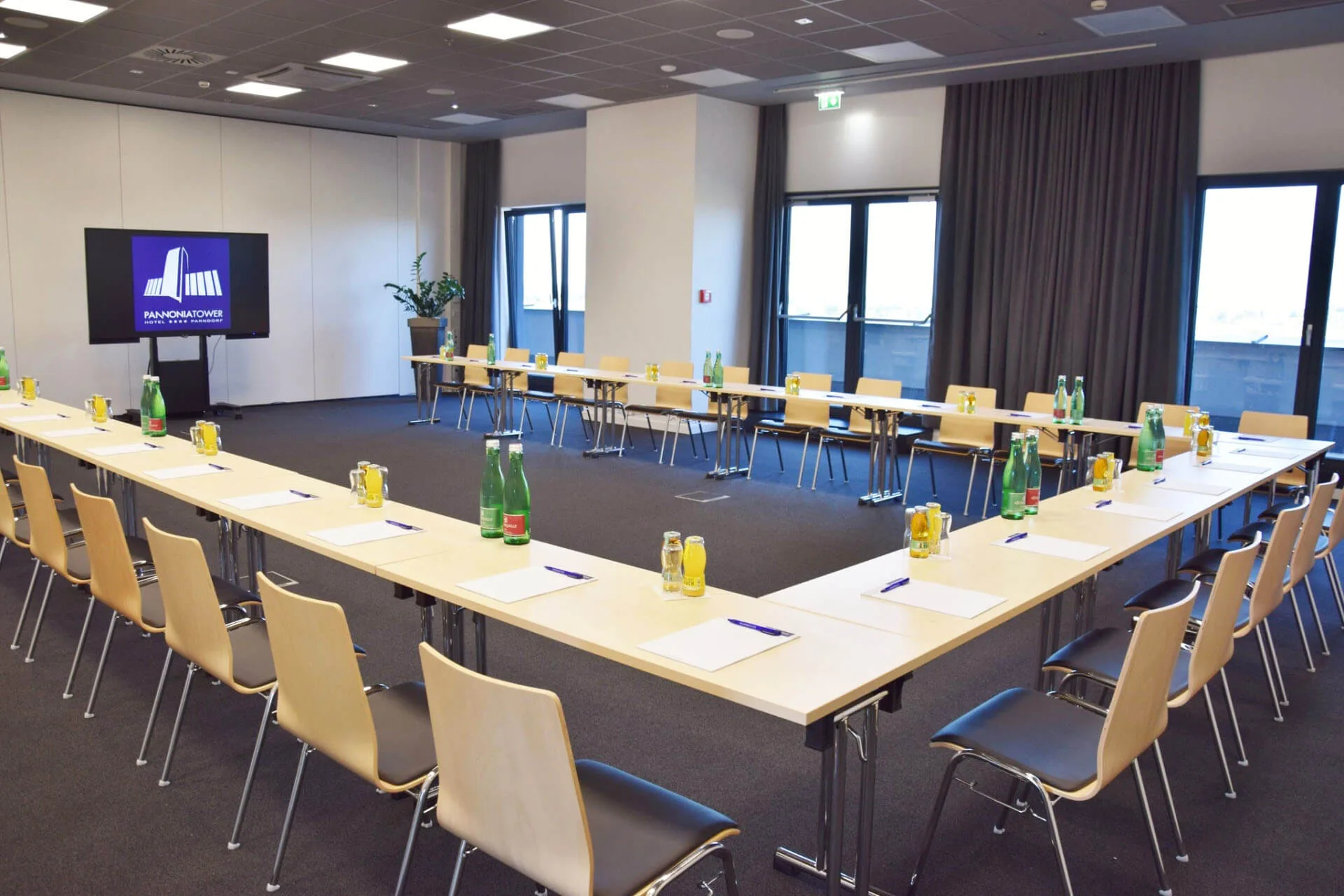 U-förmiger Konferenztisch in einem modernen Konferenzraum mit Stühlen, Wasserflaschen, Gläsern, Notizblöcken und Stiften. Im Hintergrund sind große Fenster, eine Topfpflanze und ein Bildschirm mit einem Logo zu sehen.
