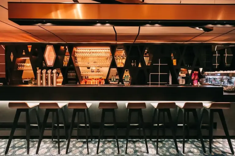 Ein modernes Barinterieur mit geometrischen schwarzen Regalen, einem langen Tresen, sechs Holzhockern und beleuchteten Regalen mit Gläsern und Flaschen. Der Boden hat ein gemustertes Fliesendesign.