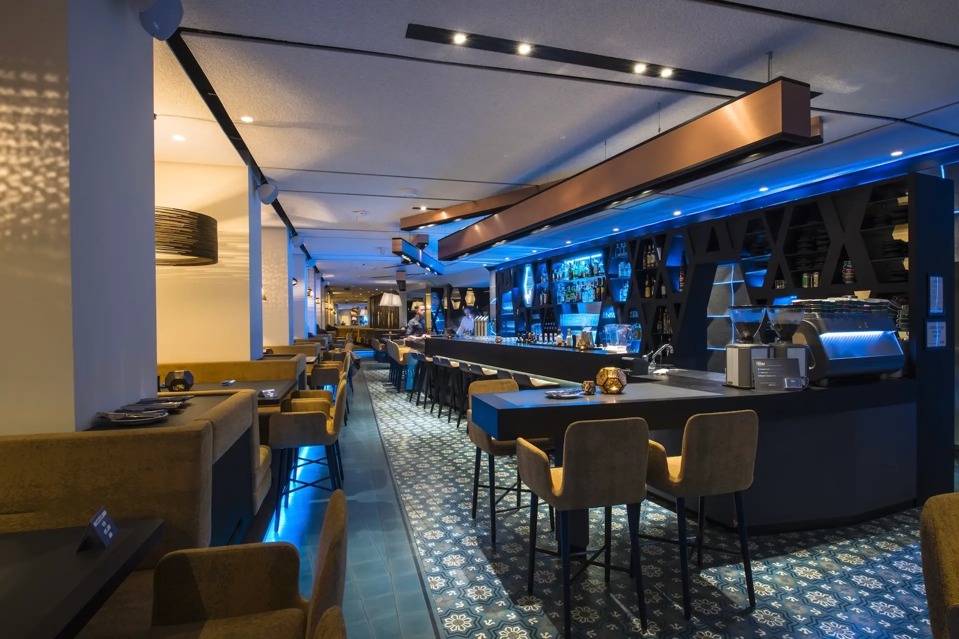 ParadisiavEine moderne, stilvolle Bar mit blauer Akzentbeleuchtung, gemustertem Kachelboden, hellbraunen Barhockern und gemütlichen Sitznischen. Die Bar ist gut bestückt und der Raum ist leer, was eine elegante, einladende Atmosphäre schafft.