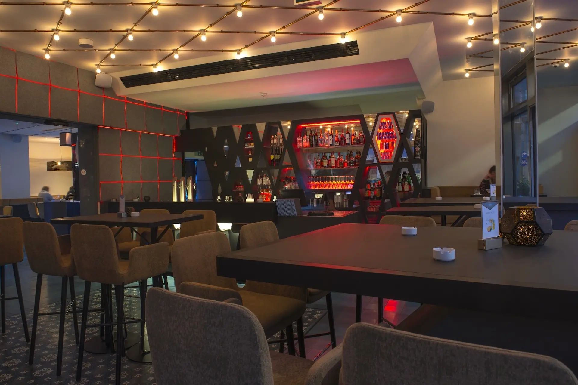 Modernes Barinterieur mit geometrischen Flaschenregalen, warmen Hängelampen und plüschigen Sitzgelegenheiten an dunklen Tischen, die eine gemütliche und stilvolle Atmosphäre schaffen.