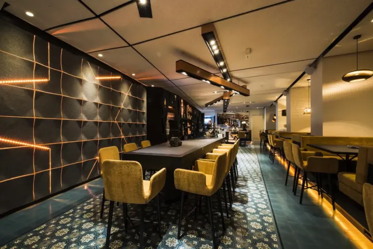 Modernes Restaurant mit gemustertem Fliesenboden, stimmungsvoller Beleuchtung, einem langen Tisch in der Mitte, umgeben von gelb gepolsterten Stühlen, geometrischer Wandgestaltung auf der linken Seite und zusätzlichen Sitznischen auf der rechten Seite.