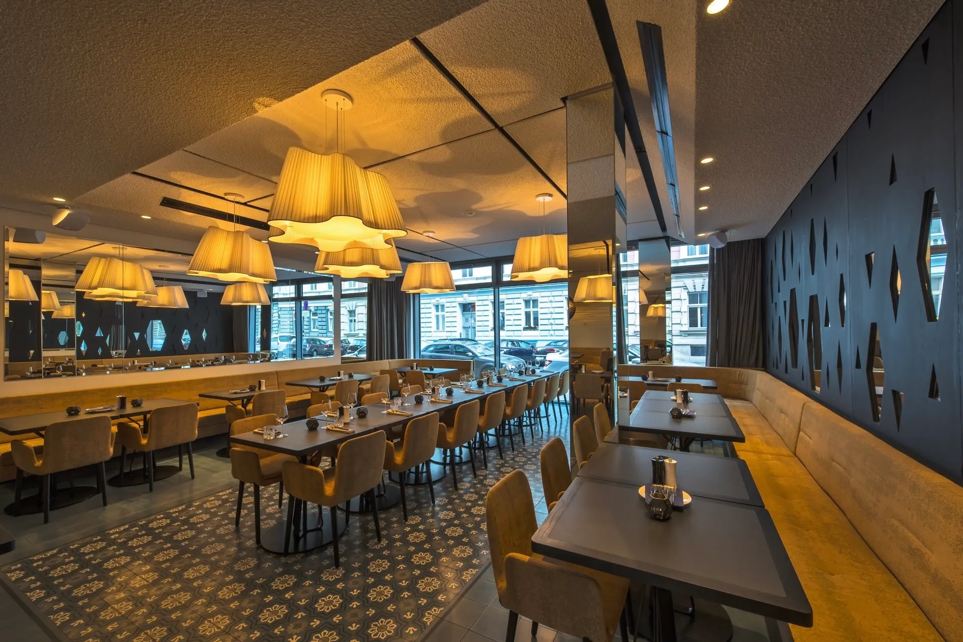 Ein modernes Restaurant mit großen Fenstern, braun gepolsterten Stühlen, Holztischen, gemusterten Fliesenböden und zahlreichen Hängelampen, die eine warme, einladende Atmosphäre schaffen.