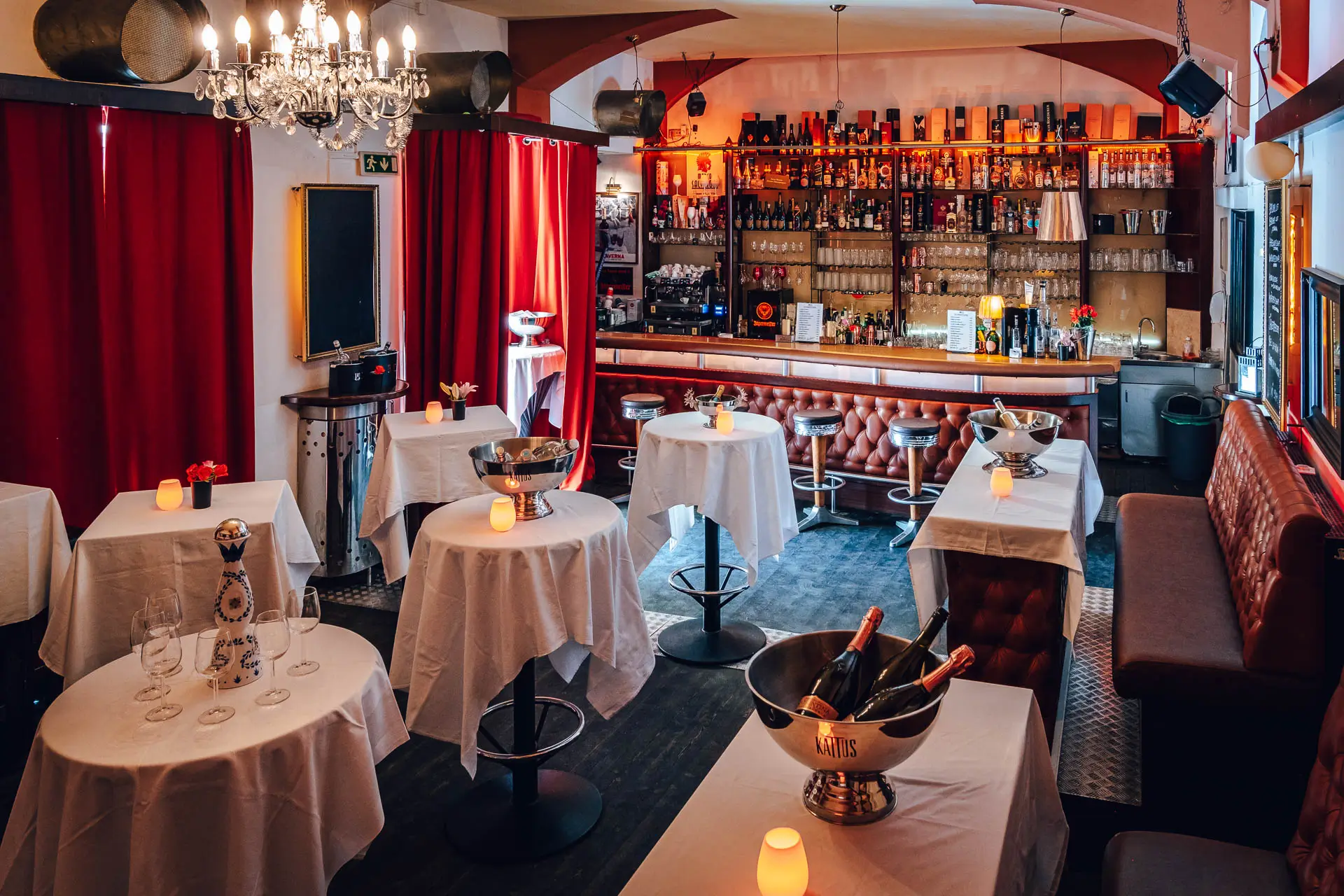Eine gemütliche Bar im Party Wien mit runden, weiß gedeckten Tischen, auf denen jeweils Eiskübel mit Sektflaschen und Kerzen stehen. Die gut gefüllte Bar, die roten Vorhänge und die warme Beleuchtung schaffen eine einladende Atmosphäre für jede Feier.