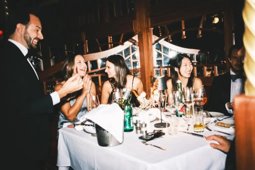 Eine Gruppe förmlich gekleideter Menschen sitzt lachend und trinkend um einen Tisch im Mirage Wien. Der Tisch ist mit Gläsern, Flaschen und Blumen in einem warm beleuchteten, eleganten Ambiente gedeckt.