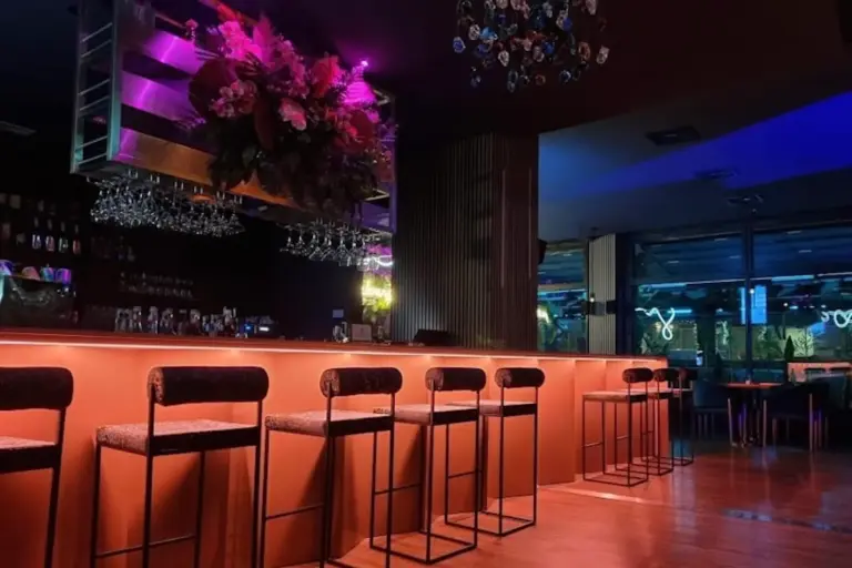 Eine moderne Bar mit leuchtend orangefarbener Unterbeleuchtung, hochlehnigen Barhockern, hängenden Weingläsern, einem großen Blumenarrangement über der Bar und farbenfroher Umgebungsbeleuchtung in einem stilvollen, schummrigen Ambiente.