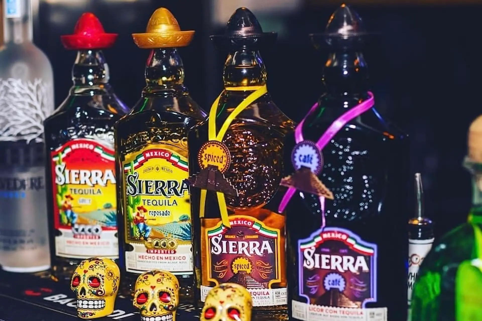 Eine Reihe bunter Sierra-Tequila-Flaschen, jede mit einem kleinen Hut auf der Kappe, steht auf einer Bar. Vor den Flaschen sind dekorative Totenköpfe angebracht, die für einen festlichen Touch sorgen. Im Hintergrund sind weitere Getränkeflaschen zu sehen.