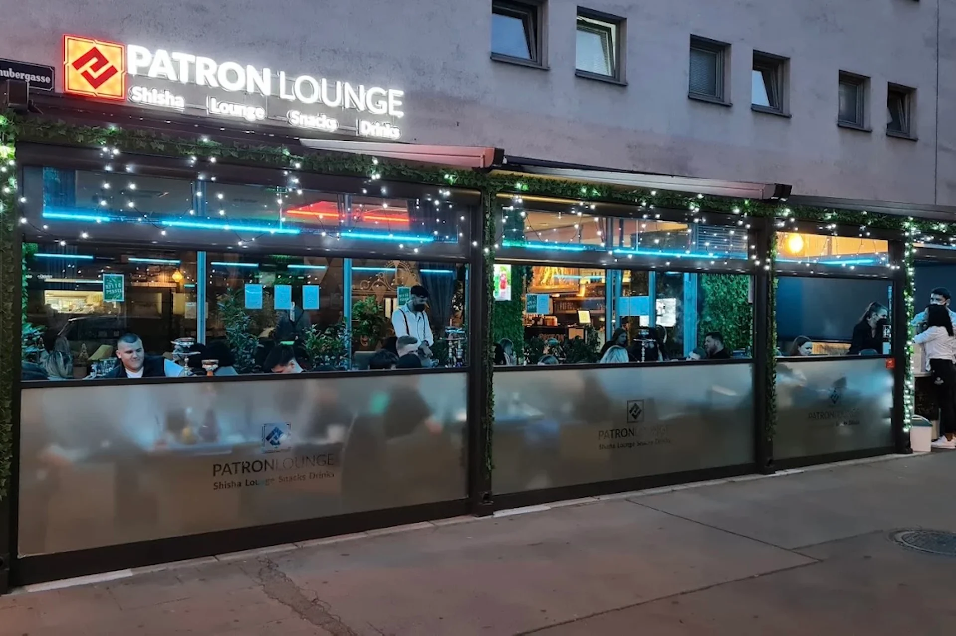 Die Fassade der Patron Lounge mit Leuchtreklamen und Glaswänden, die die Gäste im Inneren umschließen.