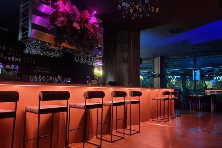 Patron Lounge Eine Bar mit Barhockern an einer orange beleuchteten Theke, über der sich ein Glasregal mit Blumenarrangements und ein bunter Kronleuchter befindet.