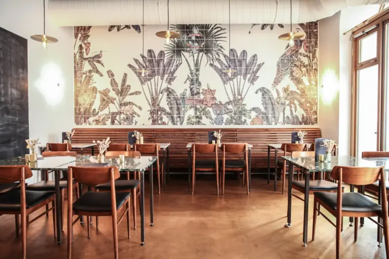 Modernes Restaurantinterieur mit Holzstühlen und -tischen, einem großen Wandgemälde mit tropischen Pflanzen und Tieren an der Wand, Hängelampen darüber und Sonnenlicht, das durch die Fenster auf der rechten Seite einfällt.