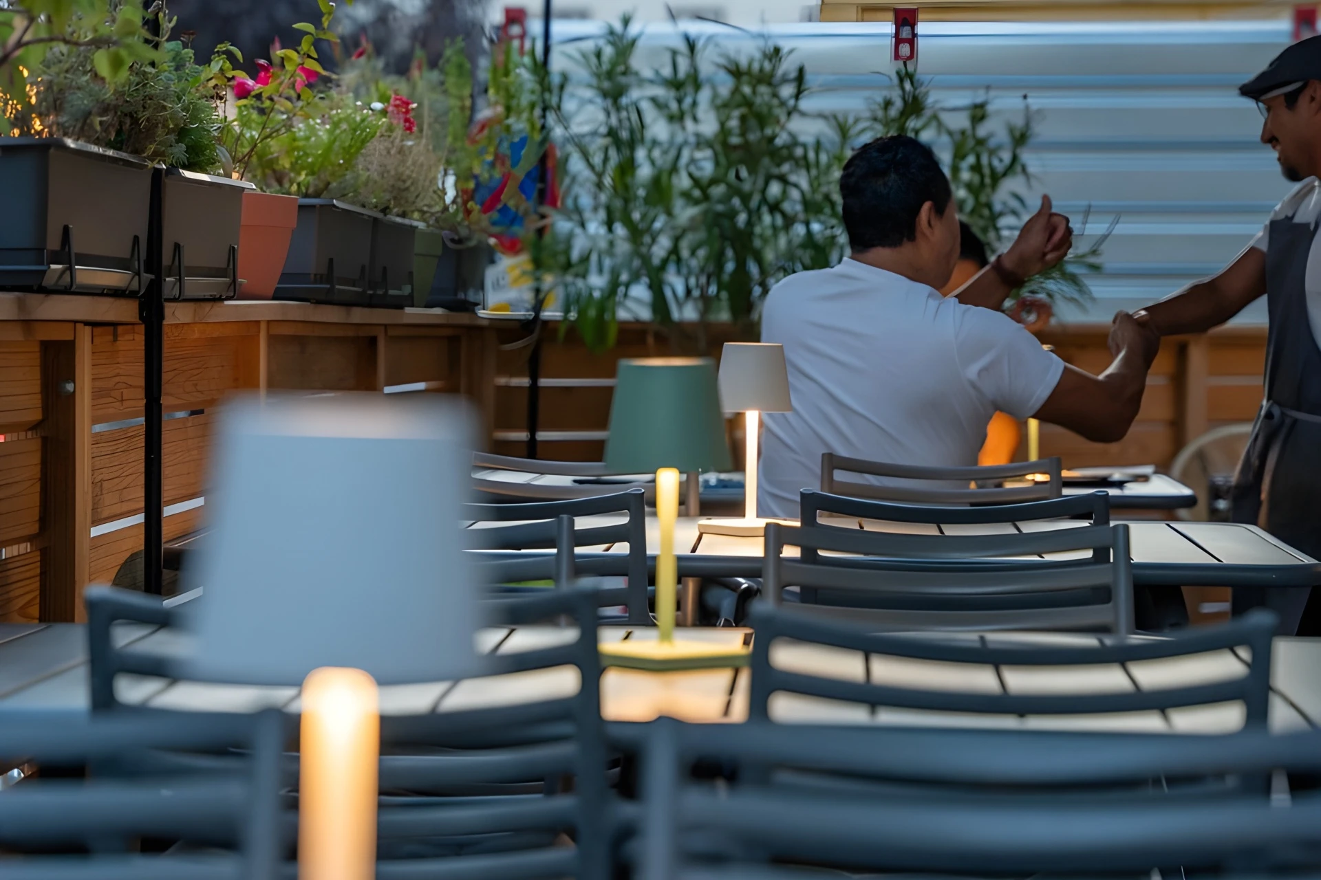 Zwei Personen unterhalten sich in einem Straßencafé mit modernen grauen Stühlen und kleinen Tischlampen. Der Bereich ist mit grünen Pflanzen und Blumen geschmückt, die eine gemütliche Atmosphäre schaffen. Der Fokus ist leicht unscharf.