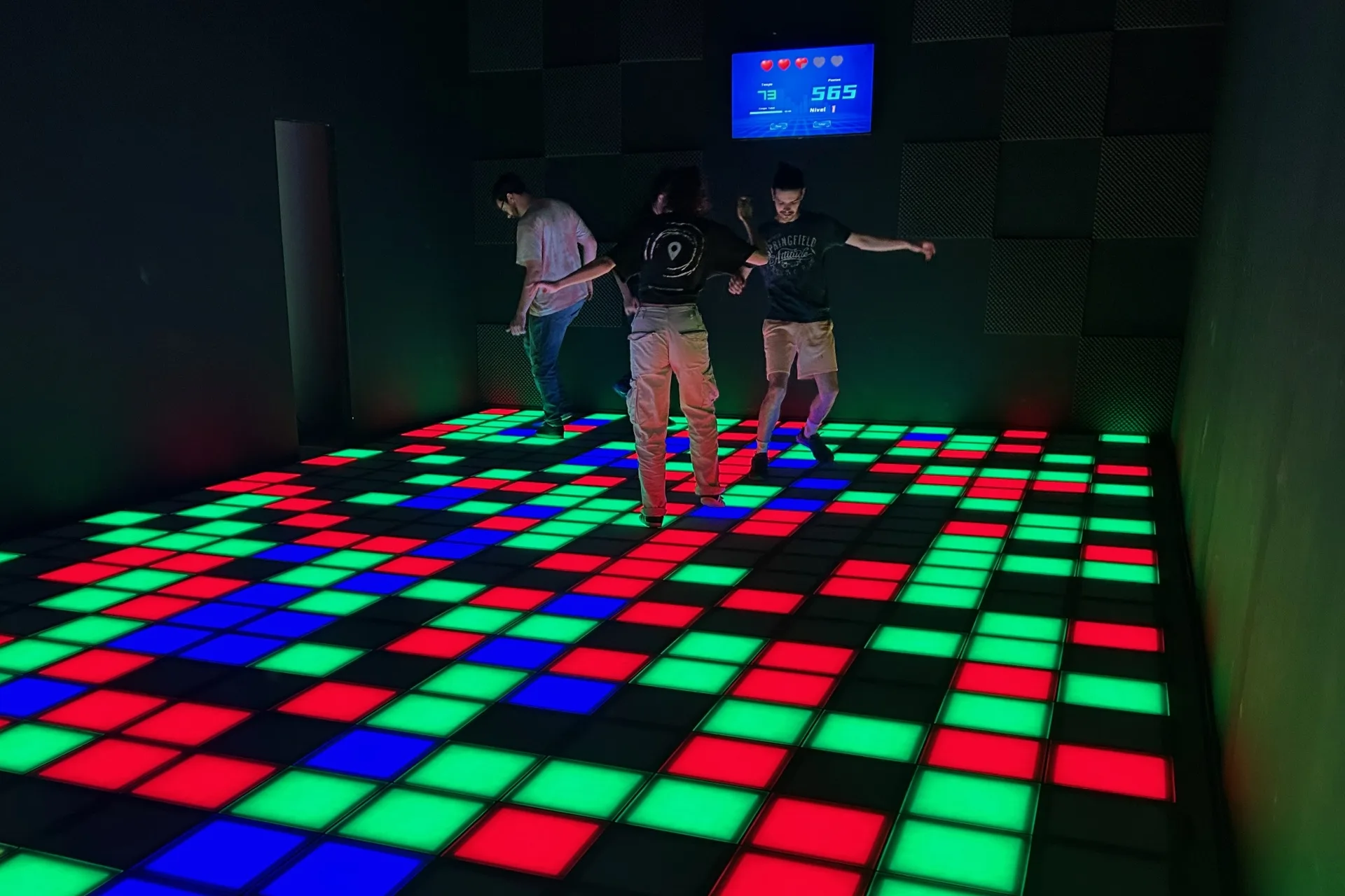 Pixel Arena Salzburg Drei Personen spielen in einem dunklen Raum auf einem beleuchteten Boden mit bunten Fliesen in Rot, Grün und Blau. An der Wand im Hintergrund hängt eine digitale Anzeigetafel, die die Punkte anzeigt.