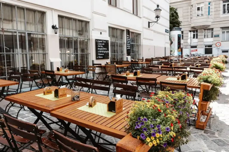 Außencafé mit Reihen von Holztischen und -stühlen auf einem gepflasterten Innenhof, der mit bunten Blumenkübeln geschmückt ist. Das Café befindet sich neben einem weißen Gebäude mit großen Fenstern und einer Speisekarte auf einer Kreidetafel.