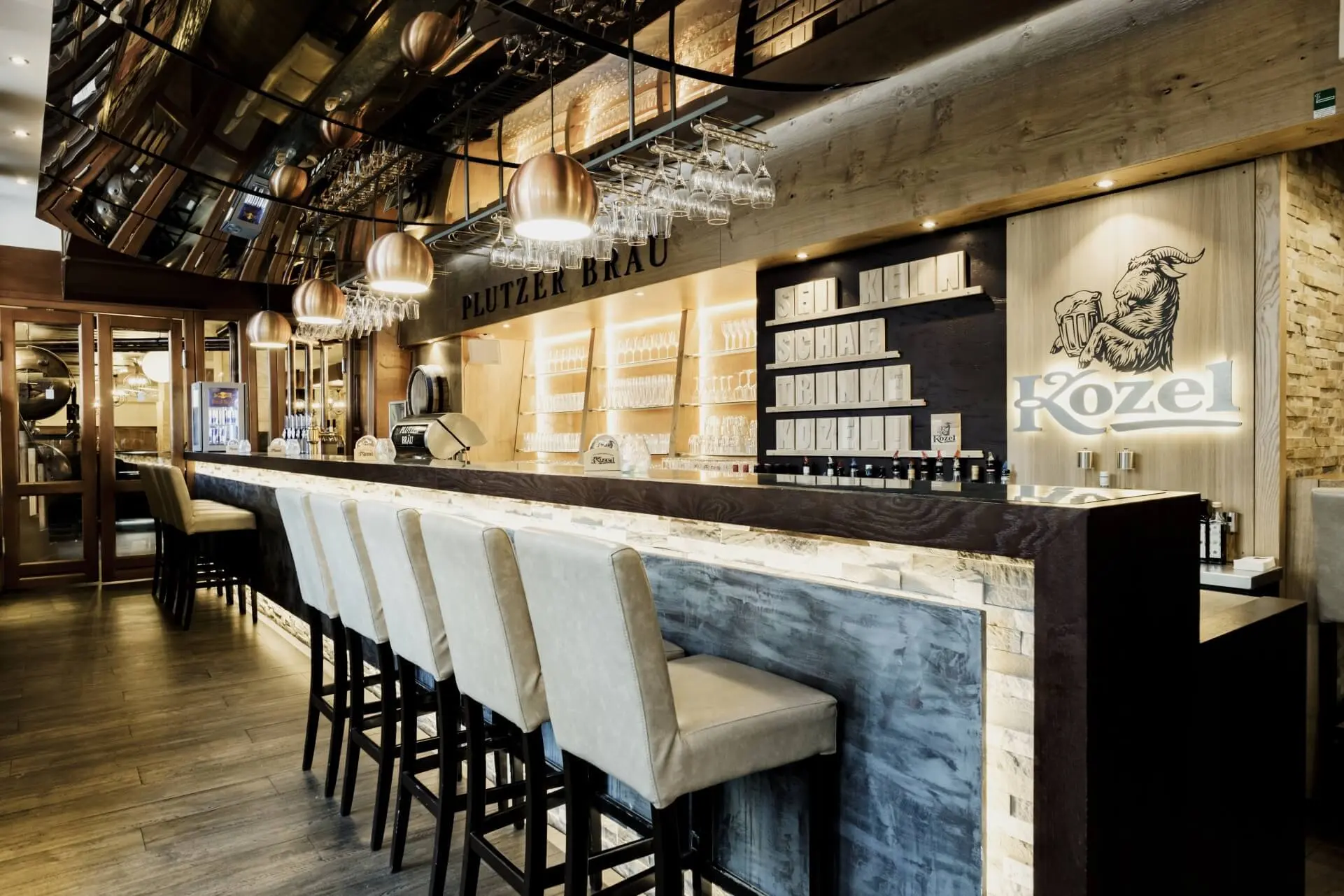 Eine moderne, stilvolle Bar mit cremefarbenen Stühlen mit hoher Rückenlehne, die an einem Holztresen aufgereiht sind, warmen Pendelleuchten, Regalen mit Gläsern und Flaschen und einem Kozel-Branding an der Rückwand.