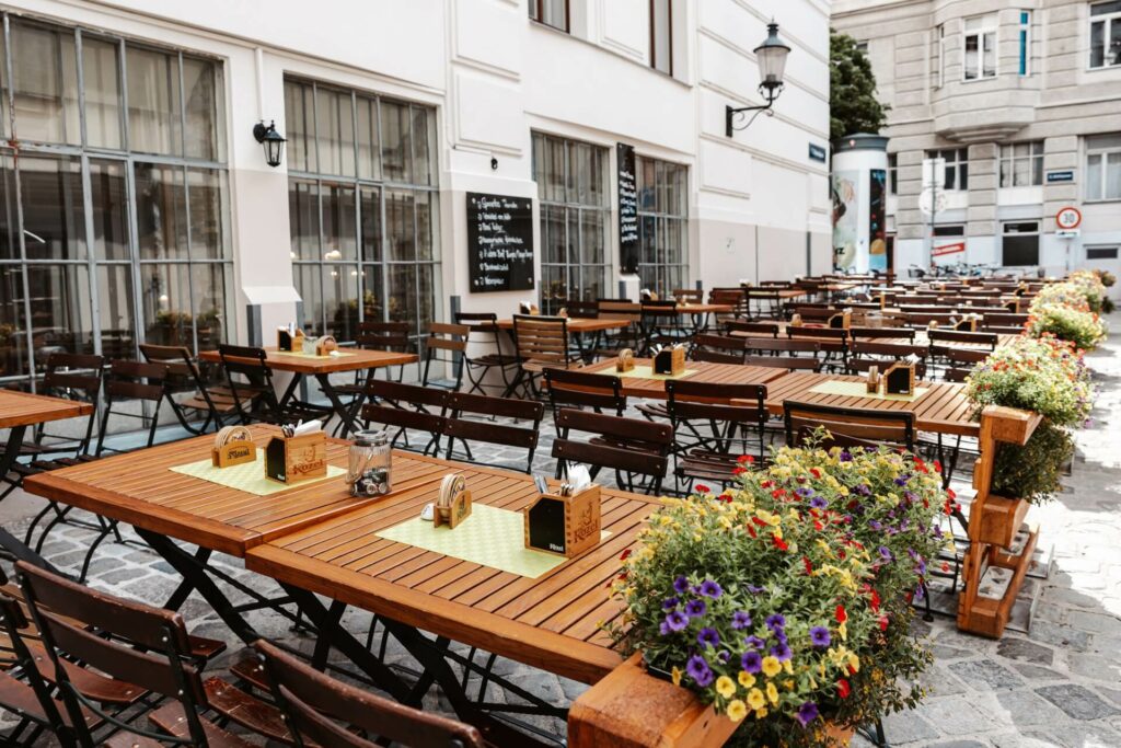 Ein Schanigarten-Café im Freien mit vielen Holztischen und -stühlen, die in Reihen auf einer kopfsteingepflasterten Straße angeordnet und mit Topfblumen an den Rändern geschmückt sind; die Umgebung wirkt leer und friedlich.