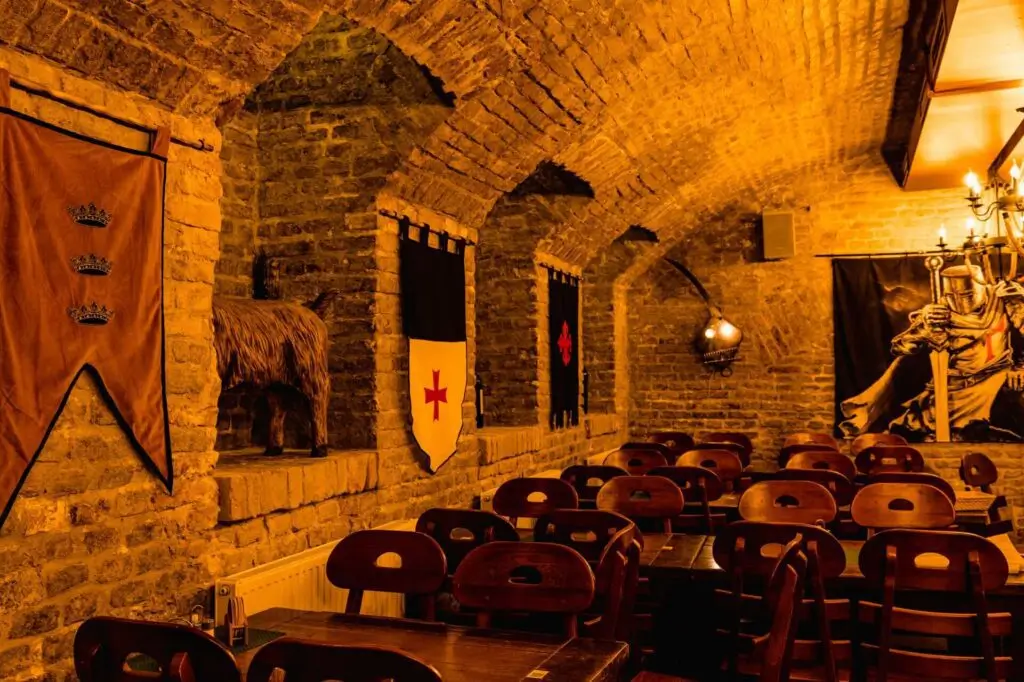 Ein Restaurant im mittelalterlichen Stil mit gewölbten Ziegeldecken, Holztischen und -stühlen, mittelalterlichen Bannern an den Wänden, warmer gelber Beleuchtung und einem Luster - perfekt für ein einzigartiges Junggesellenabschied Wien-Erlebnis.