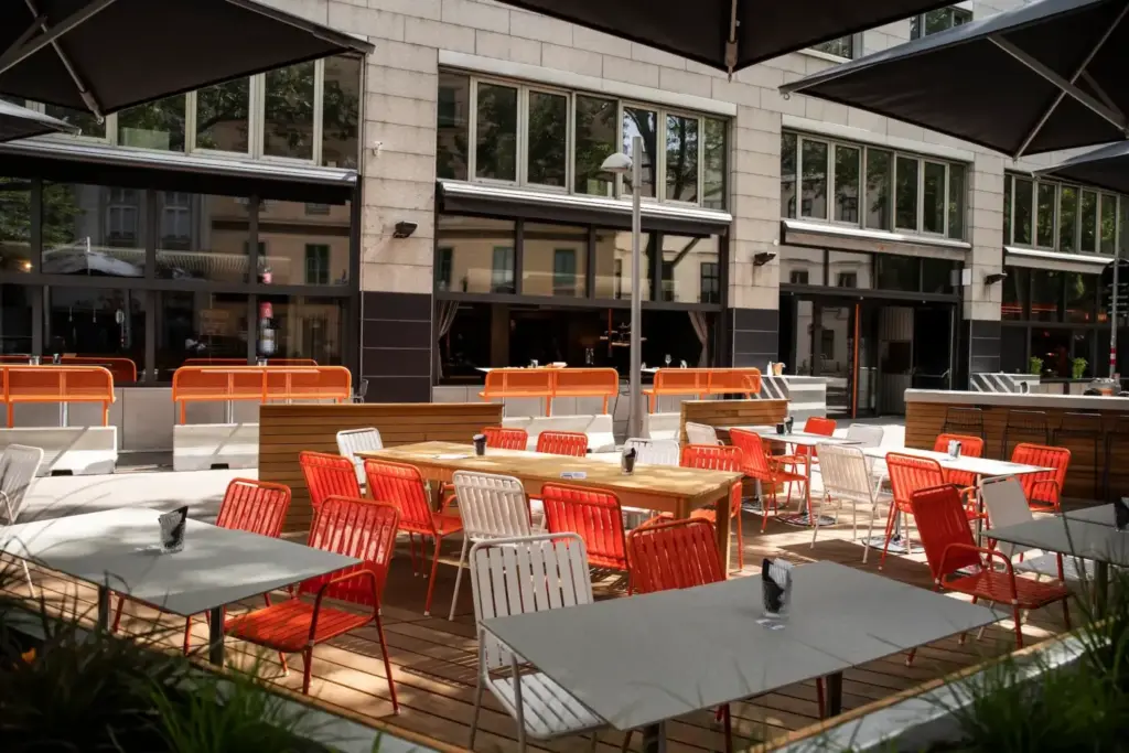 Restaurantterrasse im Freien mit Holzboden, roten und weißen Metallstühlen und rechteckigen Tischen. Große Fenster und Sonnenschirme spenden Schatten. Der leere Bereich bietet einen Blick auf den Club Praterstraße, dessen Gebäude sich in der Glasscheibe dahinter spiegeln.