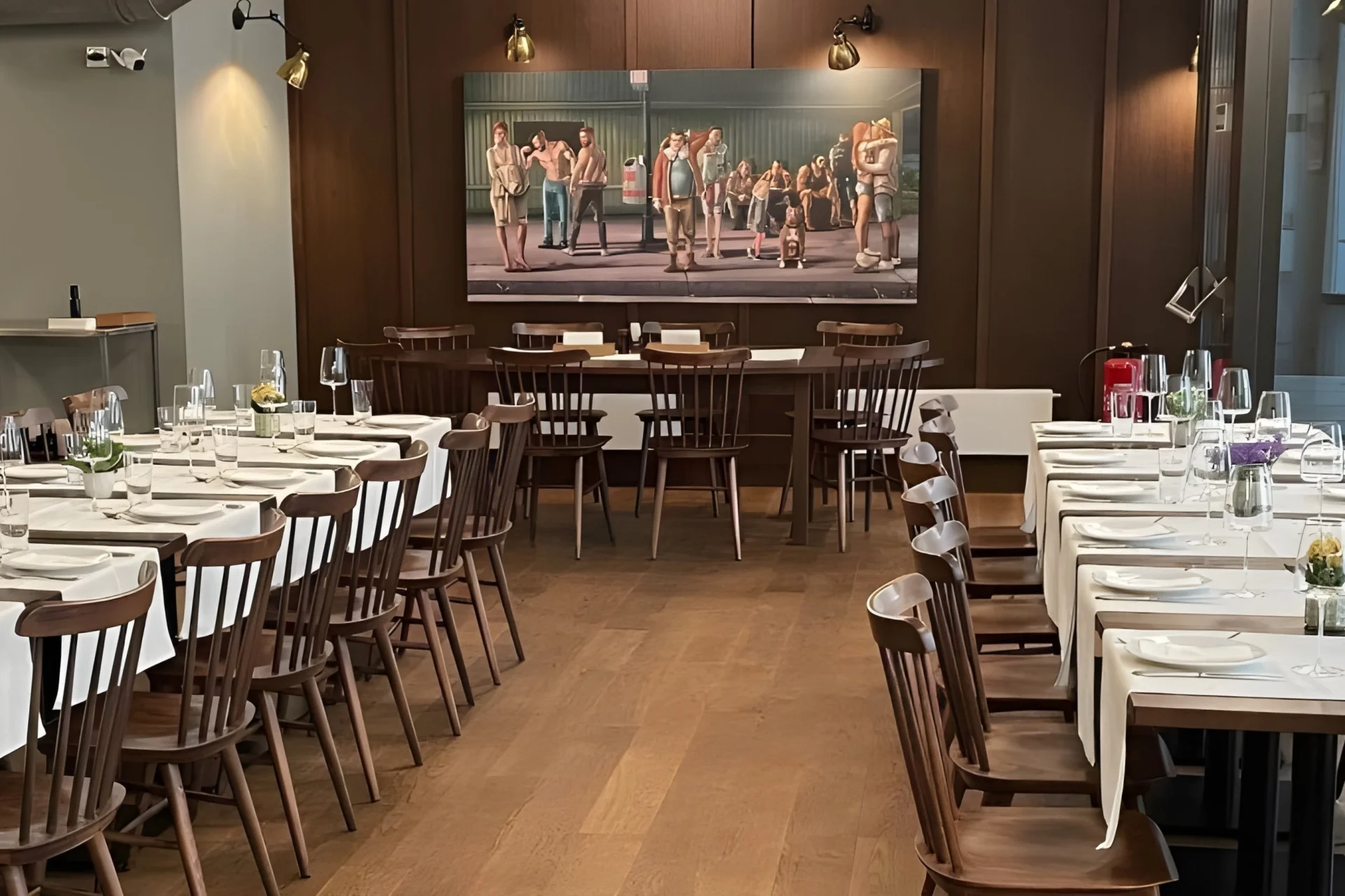 Ein ordentlich eingerichteter Restaurantbereich mit Holzstühlen und Tischen mit weißen Tischdecken, Gläsern und Besteck. An der Rückwand über einem Tisch ist ein großes Gemälde mit Menschen abgebildet.