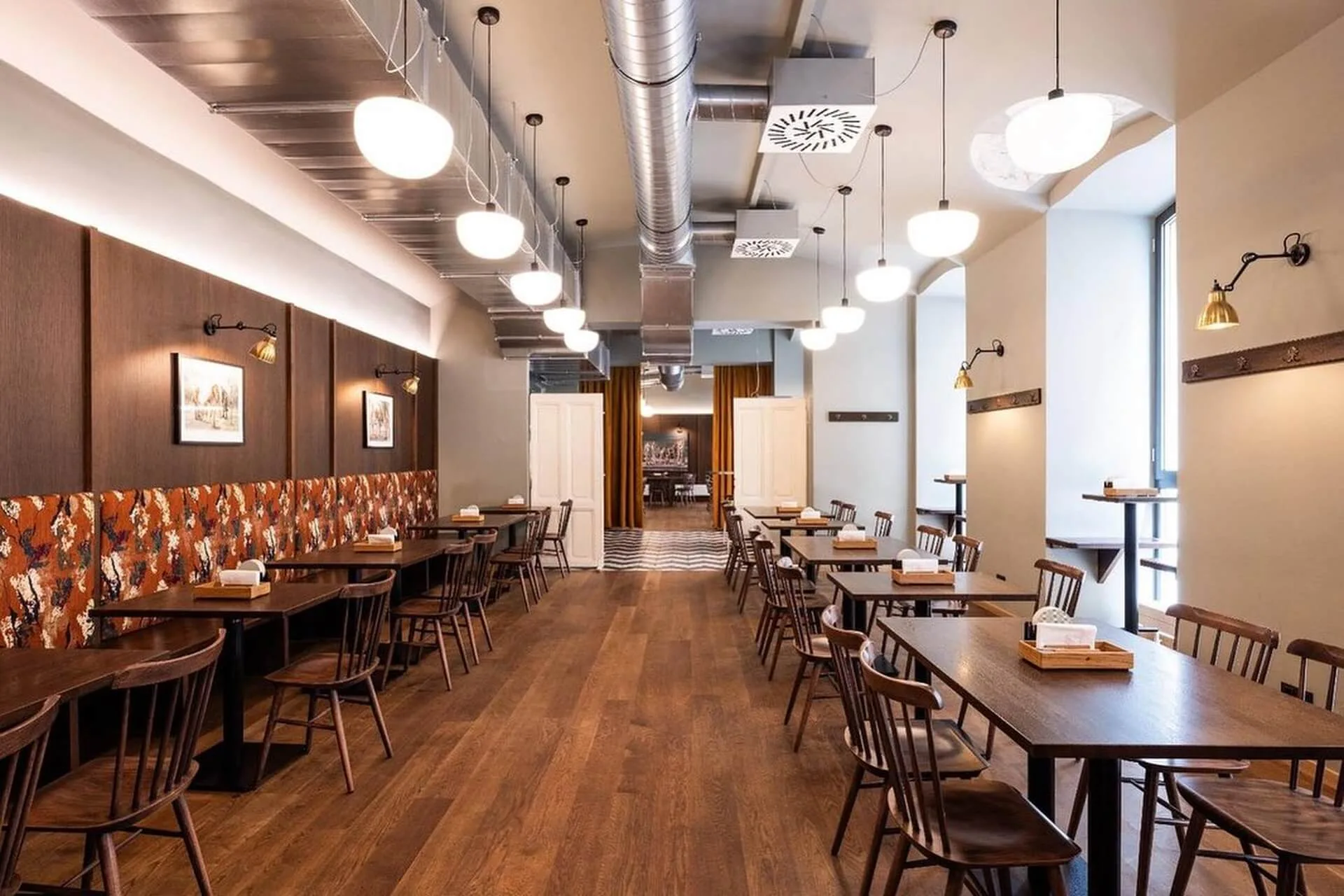 Geräumiges, modernes Restaurant mit Holztischen und -stühlen, warmer Beleuchtung, gemusterten Wandakzenten und großen Fenstern, die natürliches Licht hereinlassen. Der Raum ist aufgeräumt und unbesetzt.