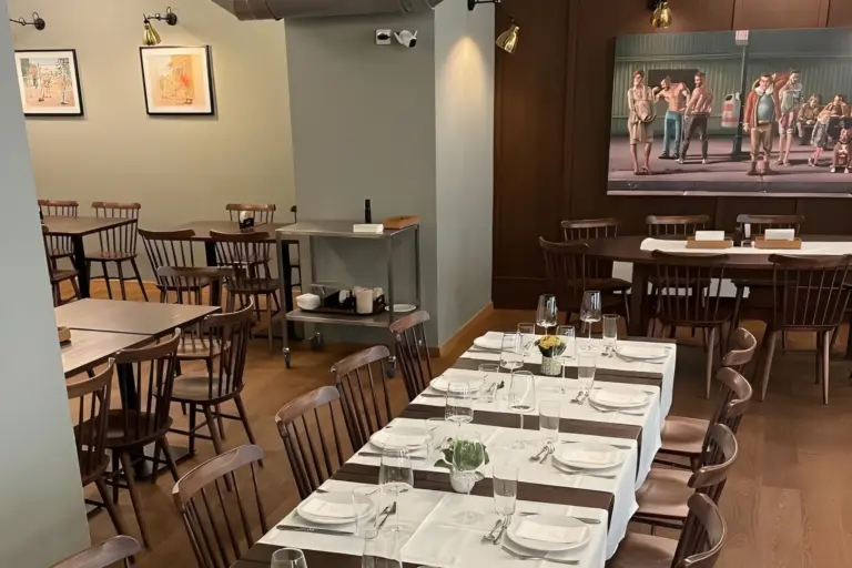 Ein Esszimmer mit Holztischen und -stühlen, einige Tische sind mit Tellern, Gläsern und Besteck gedeckt. An einer Wand steht ein Servierwagen, und im Hintergrund hängt ein großes Gemälde an einer holzgetäfelten Wand.